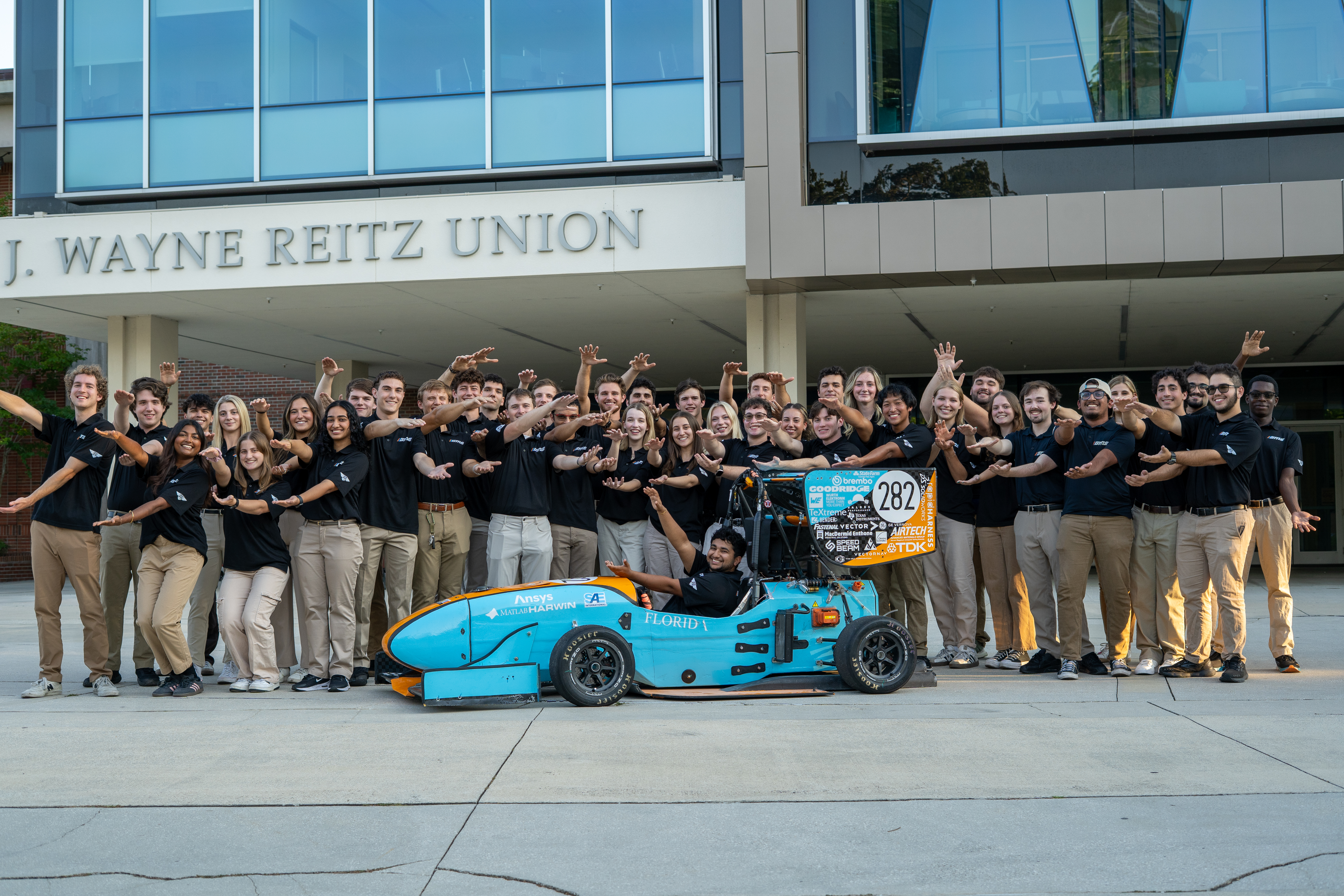 Image of the F26 team and racecar doing the gator chomp in fornt of the reitz union